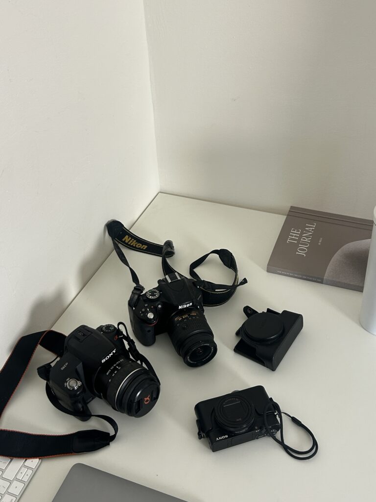 Flat lay of a clean white desk featuring Nikon and Sony cameras, a lens case, a laptop, and a journal, ideal for showcasing tools used by photographers, creatives, or virtual assistants who support visual brands.